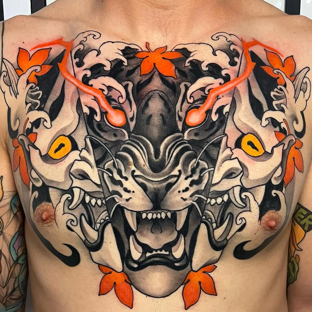 Large symmetrical tiger face chest tattoo featuring black shading, orange and yellow floral accents, ornamental scrollwork, and bared fangs.