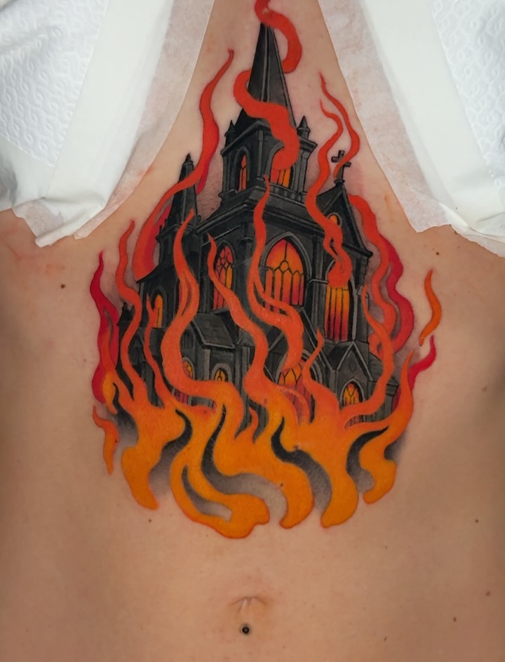 Burning church with tall spires surrounded by red, orange, and yellow flames in a teardrop shape, tattooed on upper back.