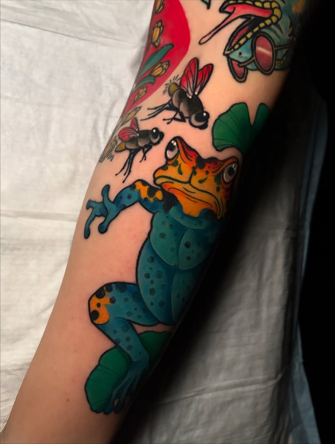 Colorful arm tattoo featuring a spotted lizard with orange and blue markings, surrounded by insects and foliage in green and red tones.