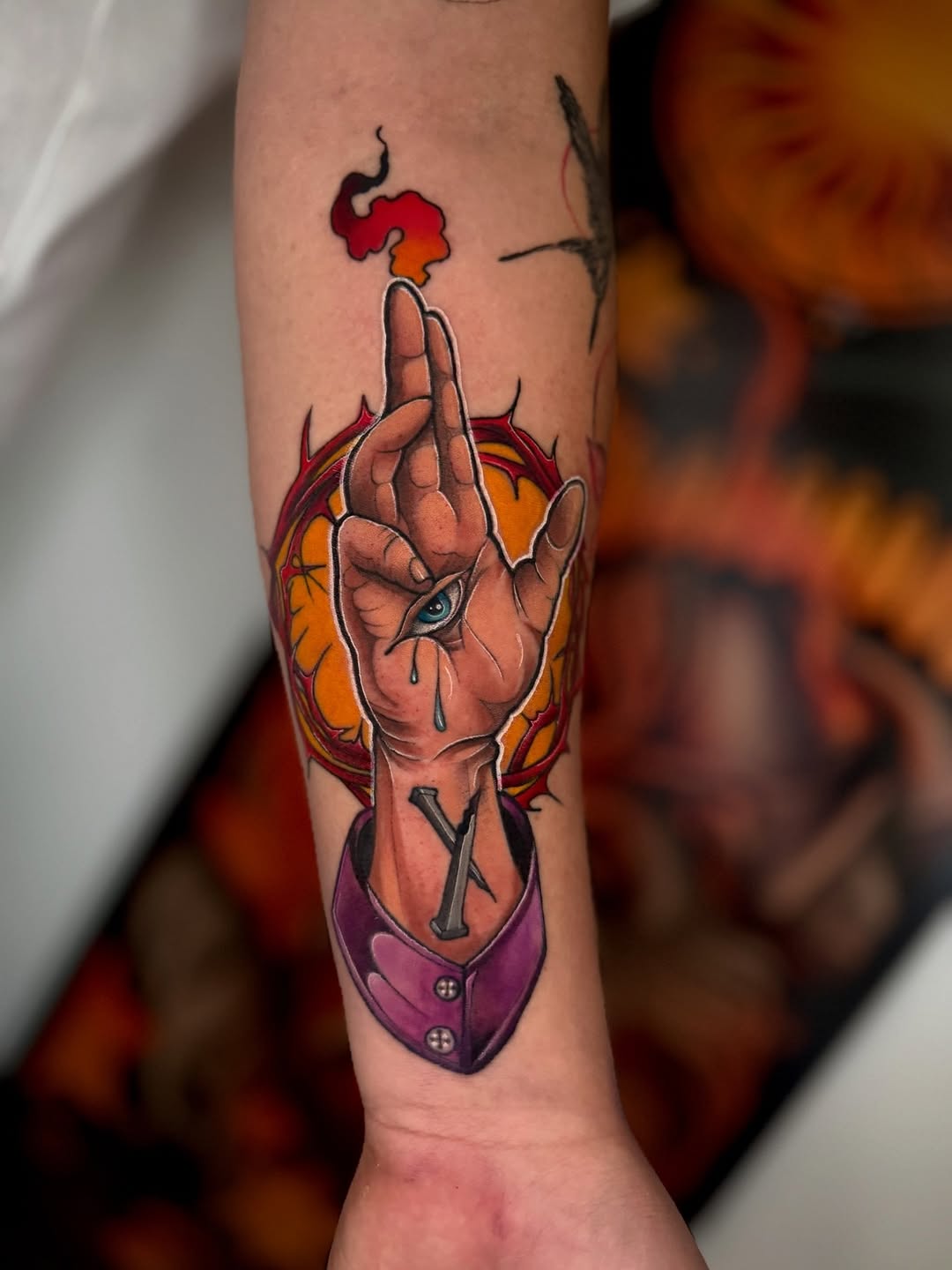 Colorful forearm tattoo of an anatomical heart with a hand gesture, flames, and a purple pouch below, rendered in red, orange, and yellow tones.