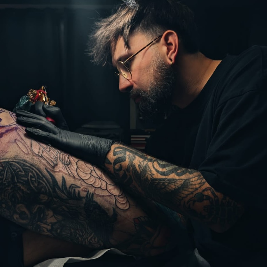 Tattoo artist wearing glasses and black gloves applies intricate dark ink designs to a client's tattooed arm during a session.