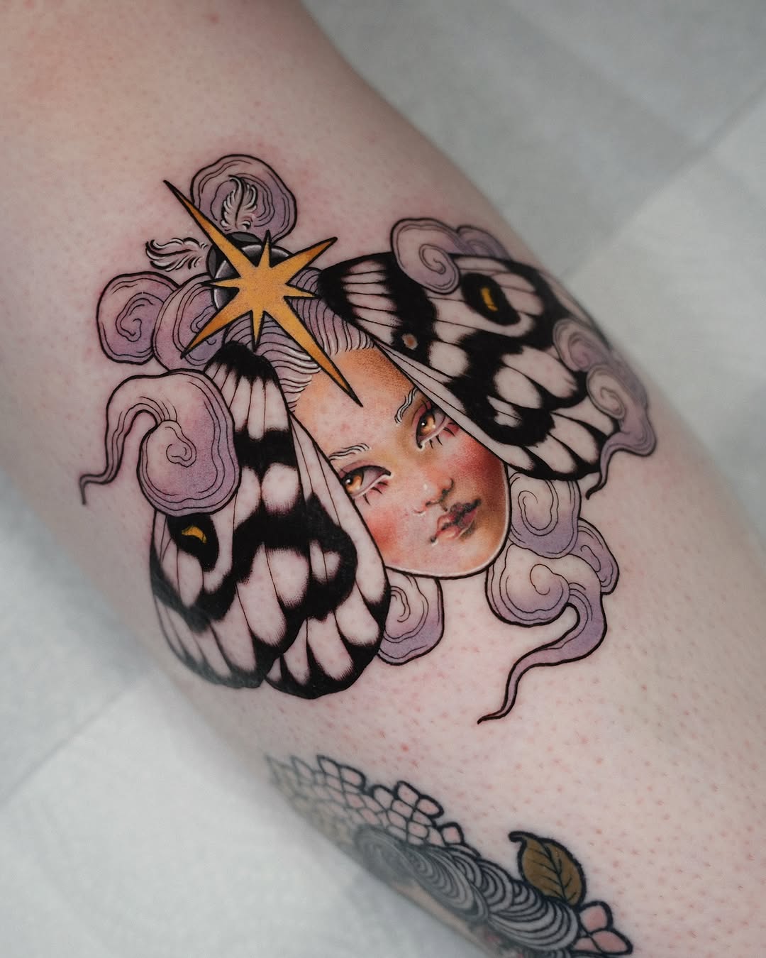 Colorful tattoo on an arm featuring a woman's face with butterfly wings, purple clouds, a gold star, and decorative swirls.