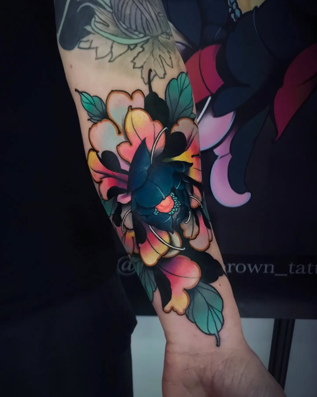Colorful floral tattoo on the forearm featuring pink, yellow, and teal flowers with green leaves and a blue bird, displayed on pale skin.