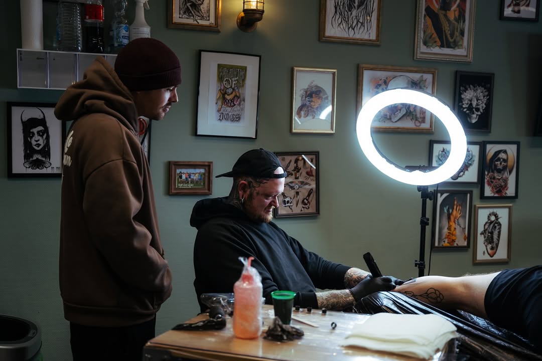 Tattoo artist with visible arm tattoos works on a client's leg under a ring light in a studio with framed artwork on sage green walls.