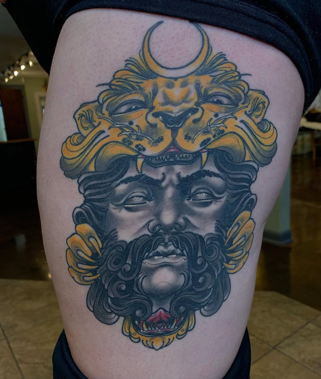 Ornate face and lion head tattoo with gold and dark blue coloring on thigh, featuring crescent moon, decorative flourishes, and red accents.