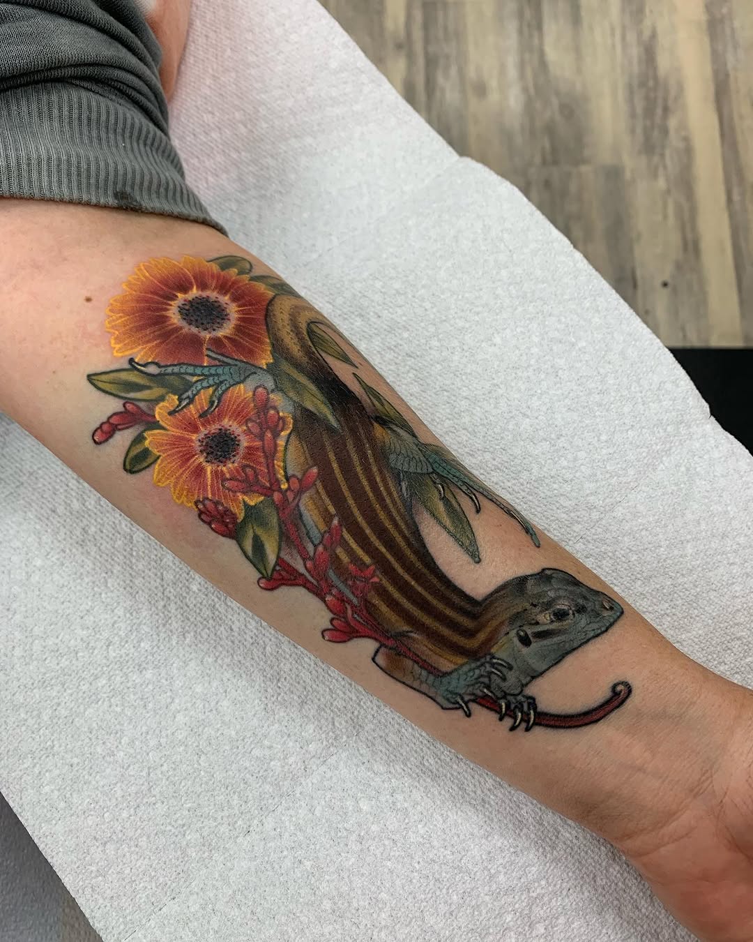 A colorful arm tattoo featuring a lizard with orange and red flowers, green leaves, and dragonfly details in vibrant yellows, browns, and teals.