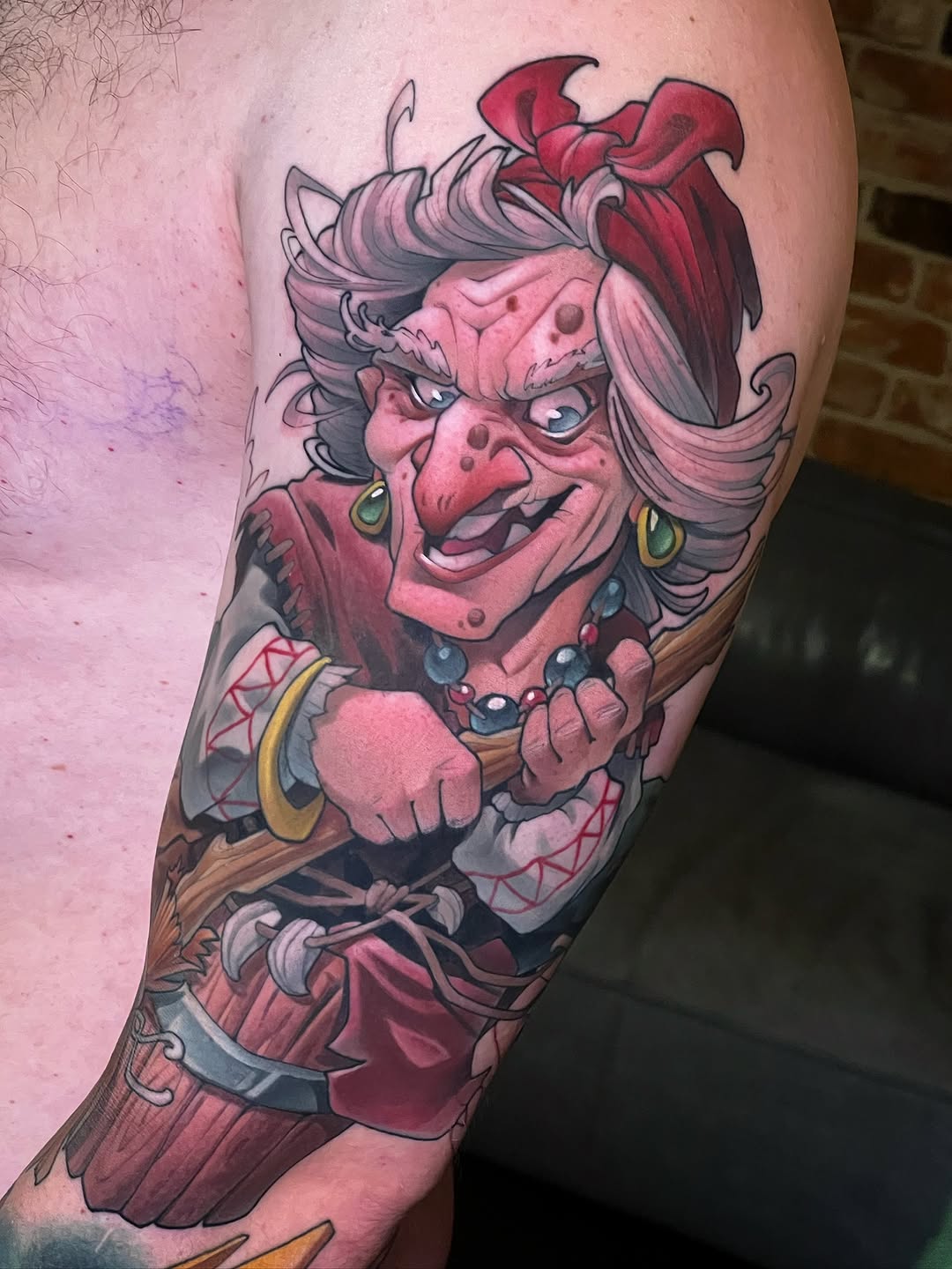 Colorful tattoo of a red-skinned character with pink hair, gold earrings, and jeweled necklace on the upper arm and shoulder.