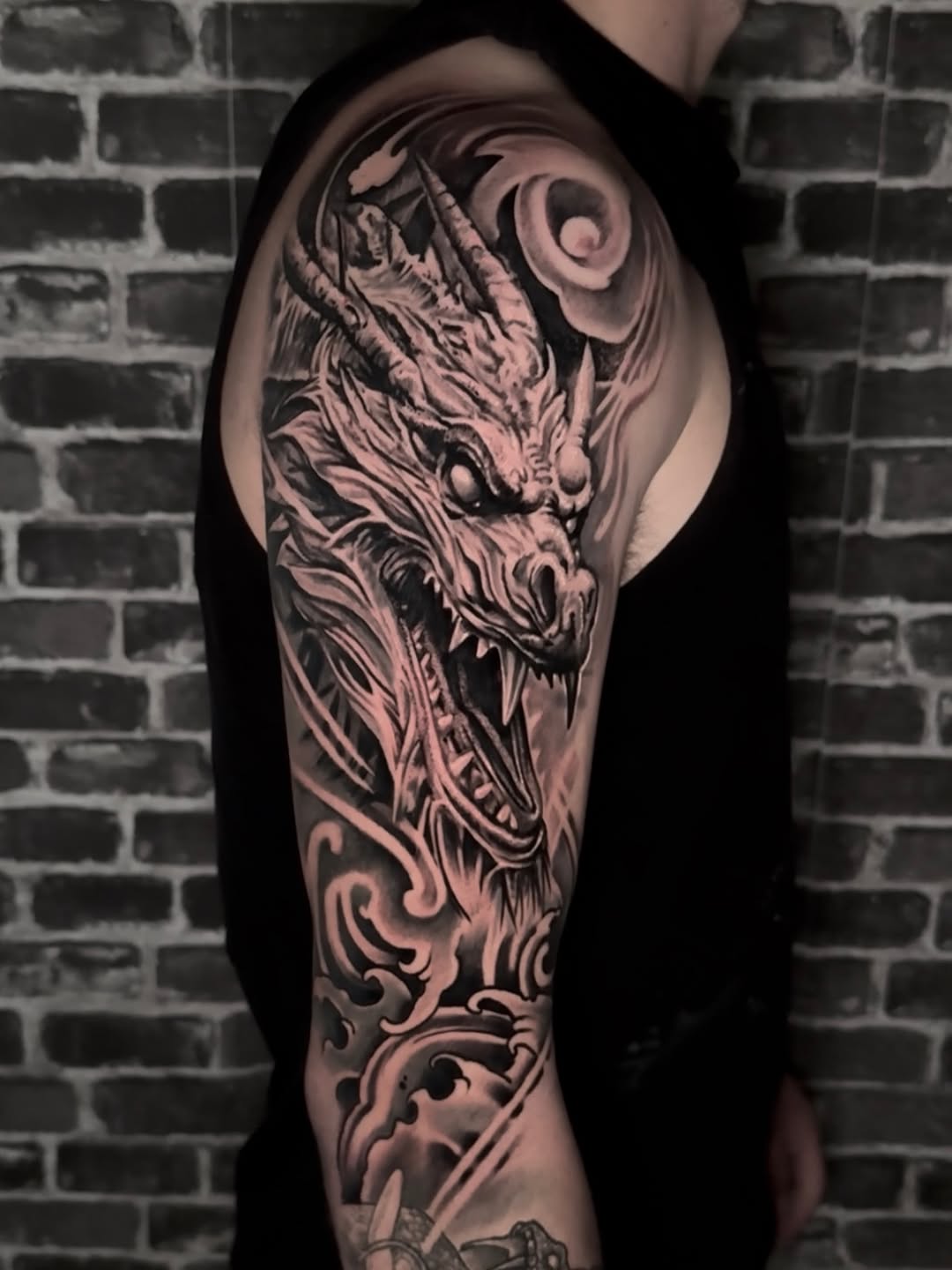 Dragon head tattoo on upper arm and shoulder with ornamental scrollwork details in black and grey ink on pale skin.