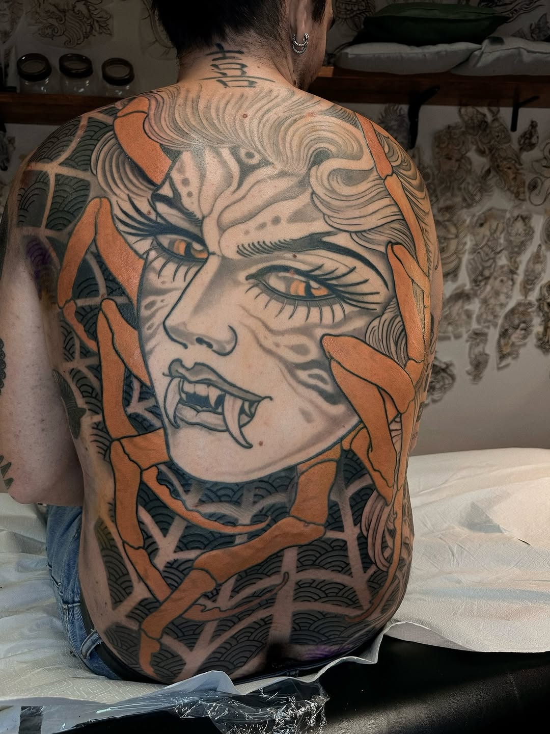 Back tattoo featuring a face with expressive eyes and open mouth, surrounded by orange petal shapes and dark patterned elements.