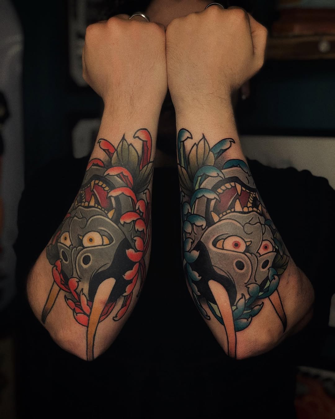 Matching hand tattoos featuring ornate masked faces with leaf motifs, colored in red, teal, and gold on both wrists.