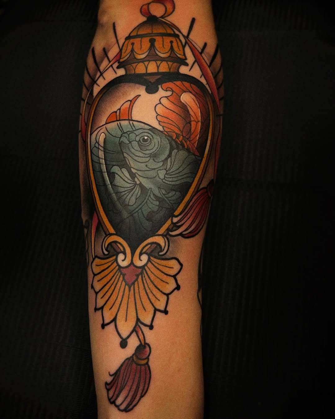 Fish in ornate lantern with decorative flourishes in orange, red, and grey ink on forearm.