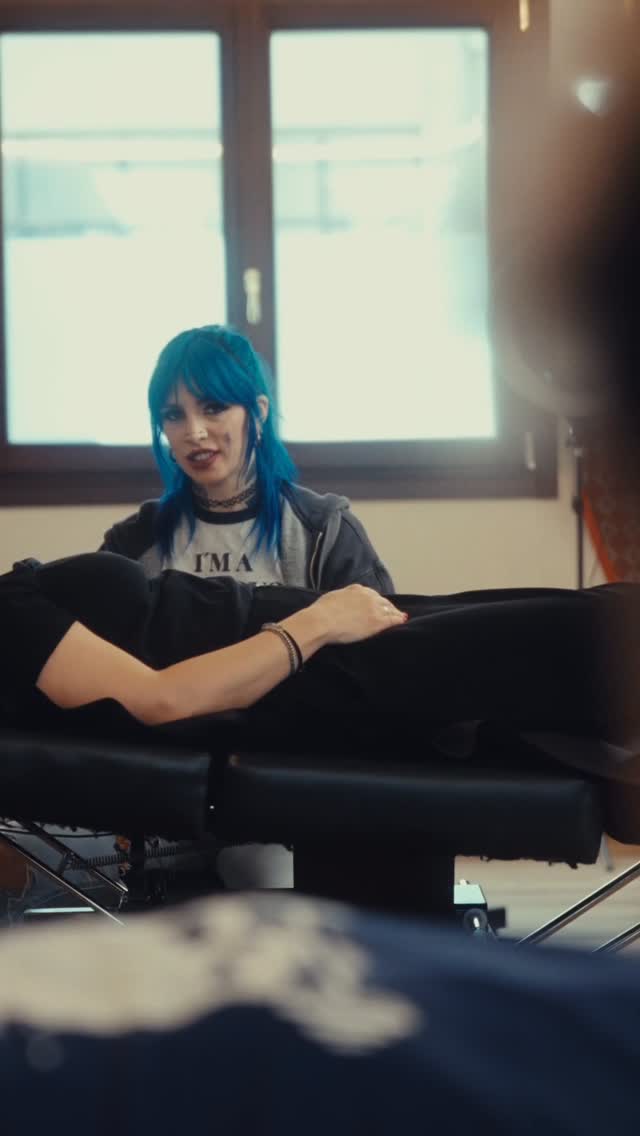Woman with blue hair wearing a black choker and graphic tee displays a wrist tattoo featuring delicate line work in black ink.