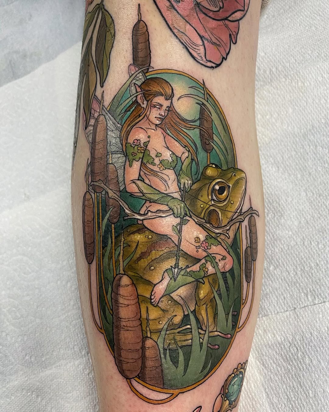 Colorful tattoo on a calf featuring a fairy with wings surrounded by frogs, cattails, and aquatic plants in greens, browns, and flesh tones.