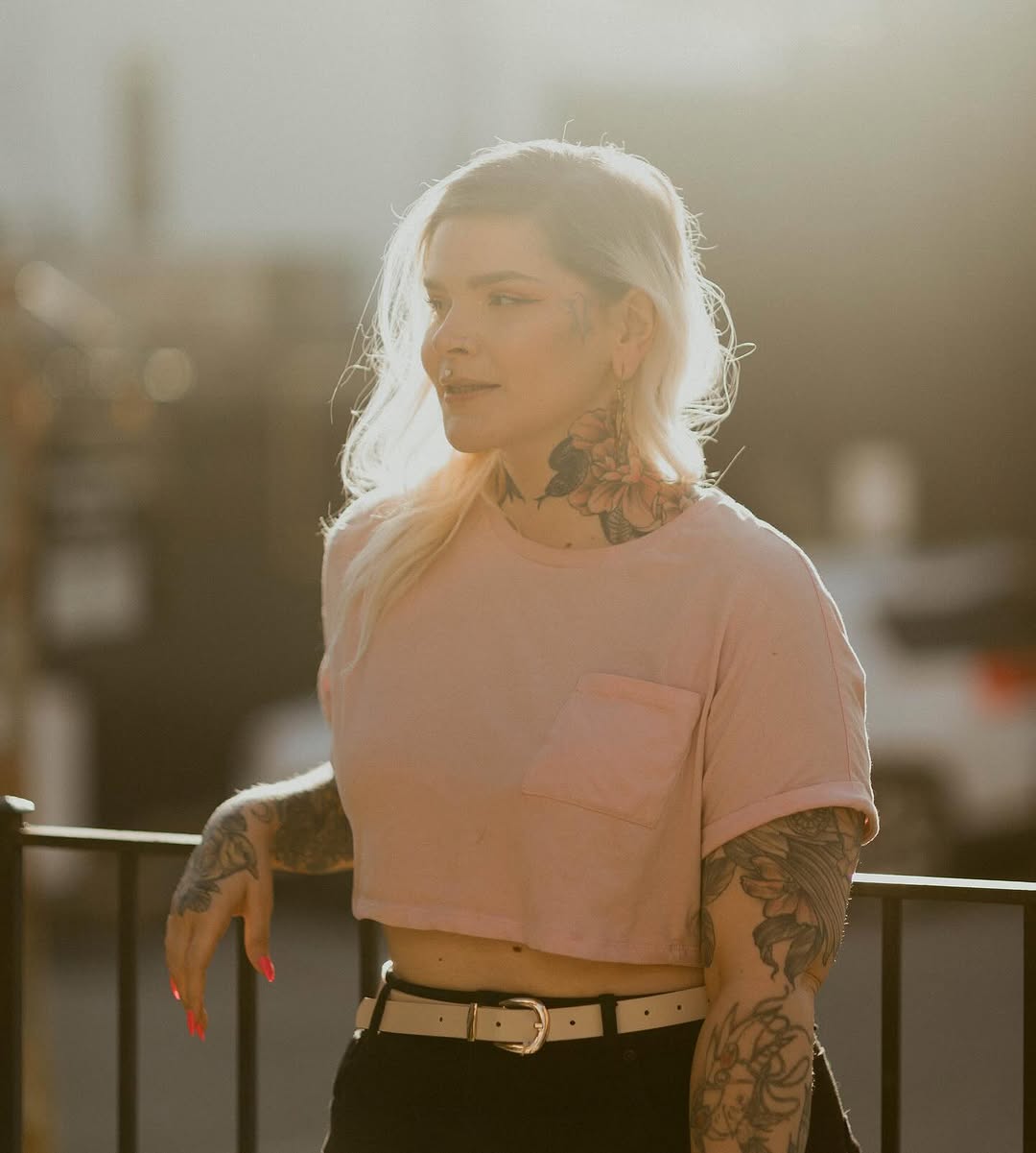 Person wearing a pink cropped shirt displaying large floral and decorative tattoos on both arms and neck in dark ink.
