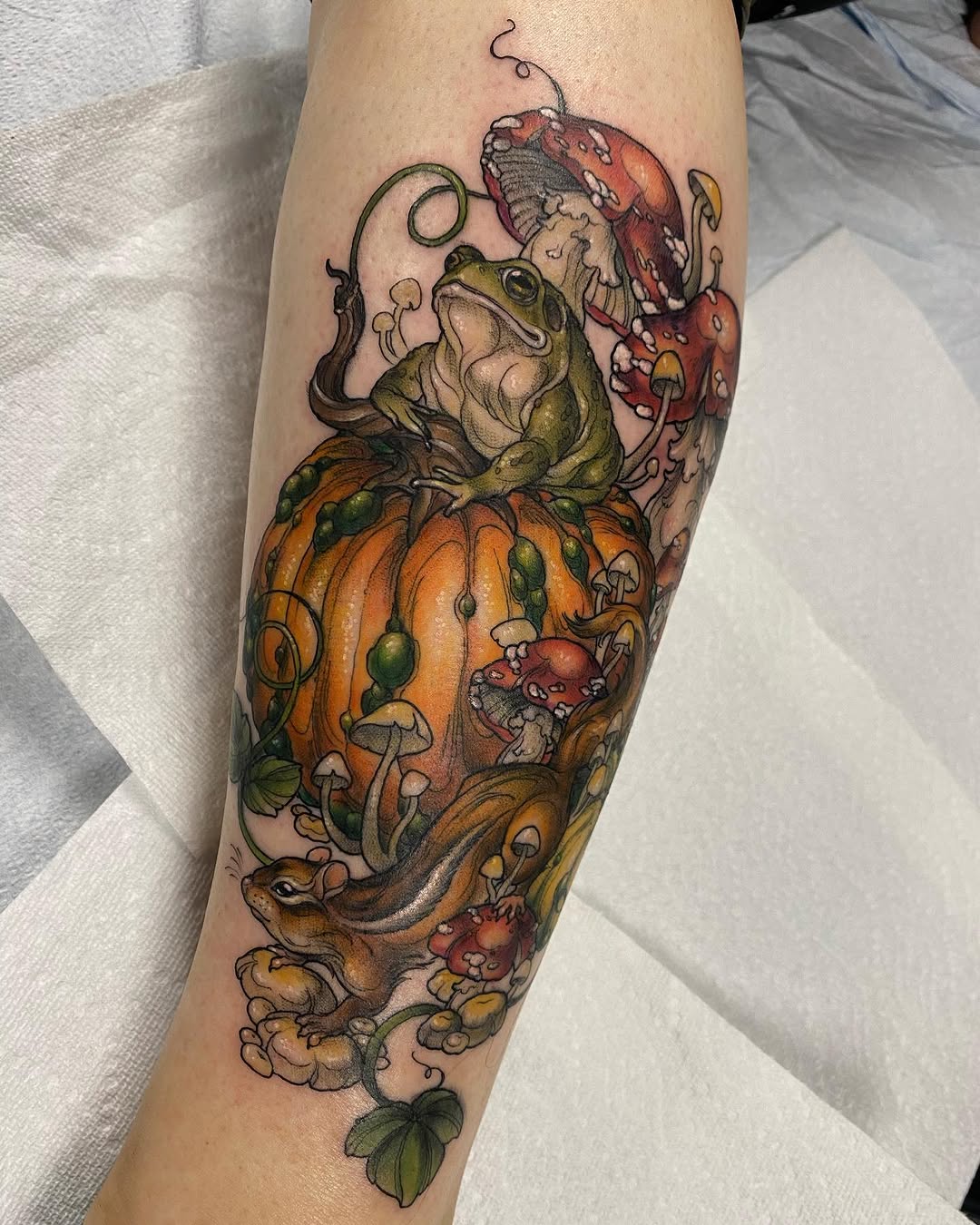 Colorful tattoo on forearm featuring a toad on an orange pumpkin surrounded by mushrooms, toadstools, vines, and small creatures in red and green tones.