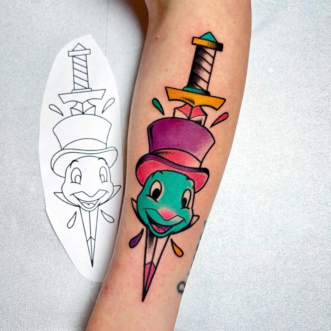 Colorful cartoon character tattoo with stacked hats and sword on upper arm, shown alongside black line drawing reference design on white paper.