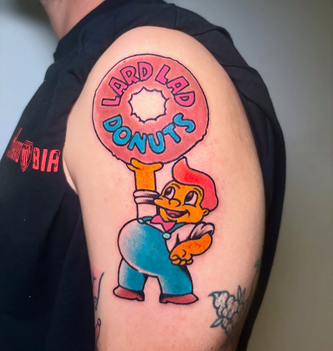 Colorful shoulder tattoo featuring a cartoon character in blue pants holding a giant red donut above their head with "Lard Lad Donuts" text.