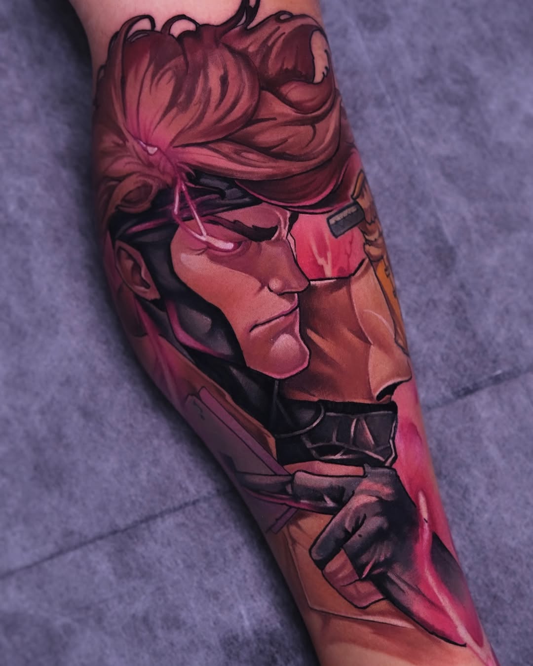 Colorful tattoo on forearm featuring a profile portrait of a figure with flowing hair and detailed facial features in red, pink, and black tones.