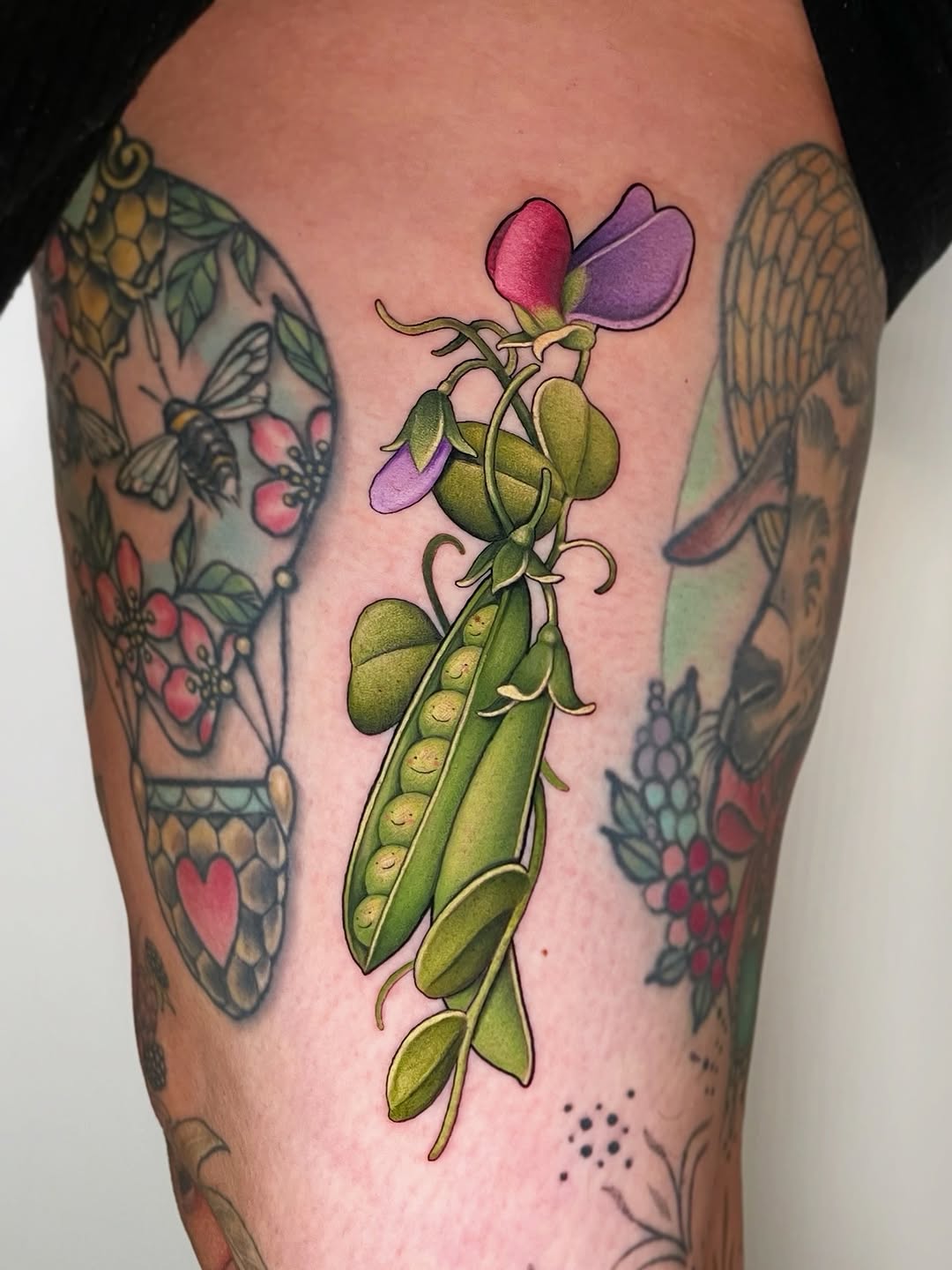 Sweet pea plant with purple and pink flowers and green pods tattooed on thigh, surrounded by other botanical designs featuring berries and butterflies.