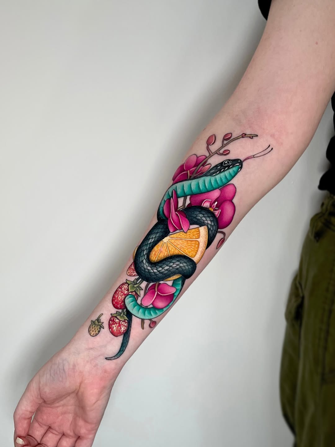 Colorful arm tattoo featuring a black snake coiled around pink flowers, an orange slice, and strawberries, with a teal fish above.