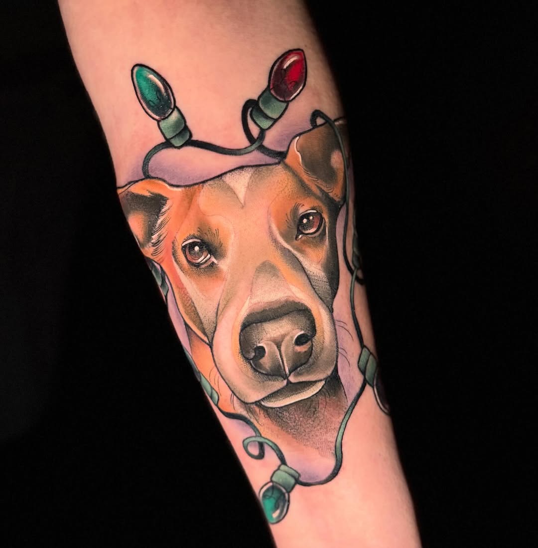 Dog's head with string lights wrapped around it featuring green and red bulbs, tattooed on the forearm in orange, brown, and green colors.