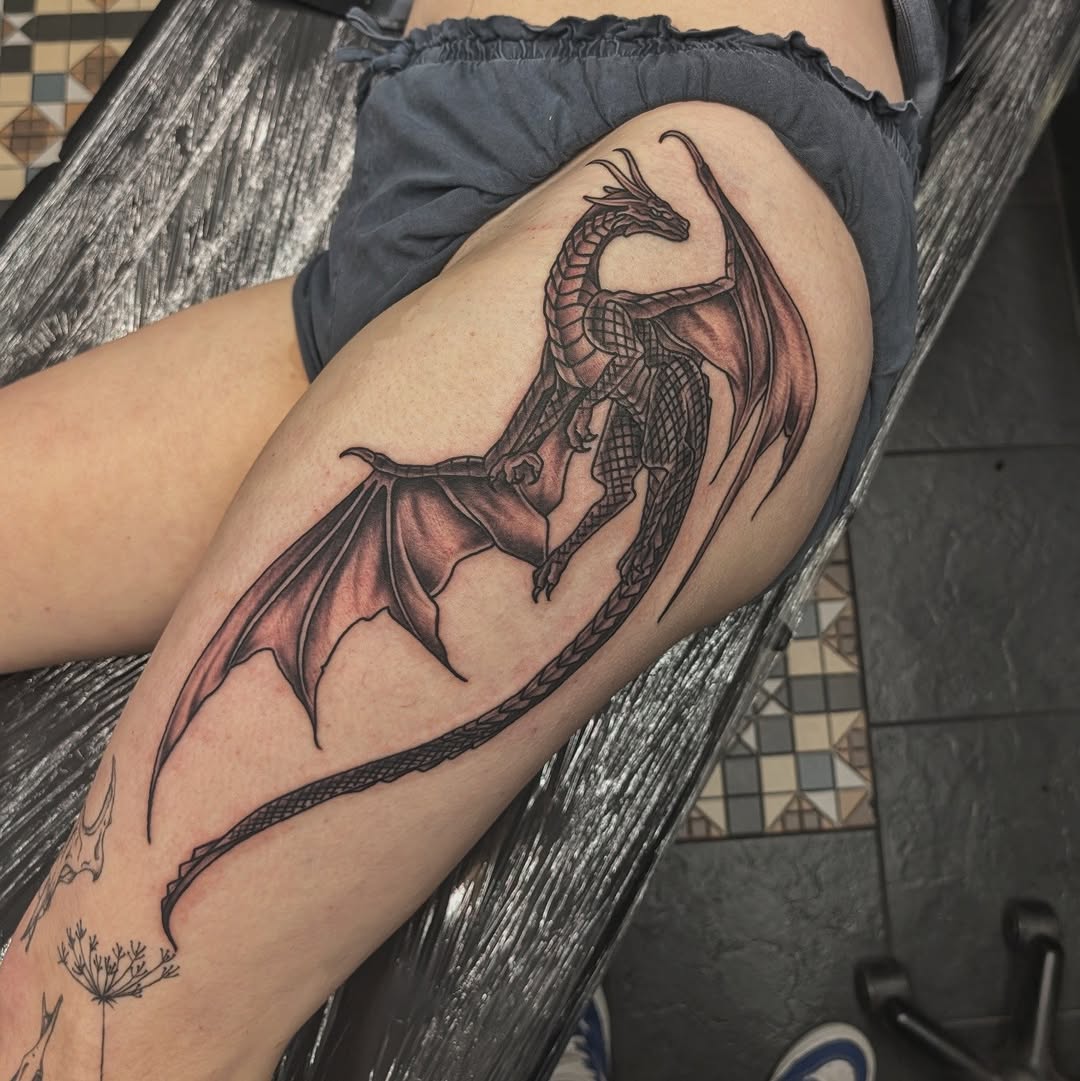 Dragon tattoo on thigh with spread wings and detailed scales in black and red ink, displayed on seated person's leg.