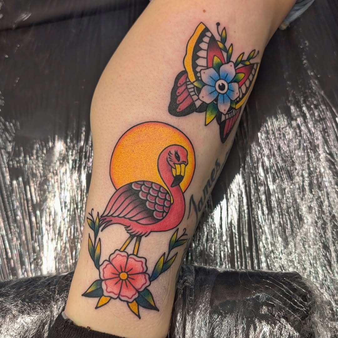 Colorful leg tattoo featuring a flamingo with orange sun backdrop, butterfly with flowers above, and pink flower below, all with vibrant orange, pink, blue, and green details.