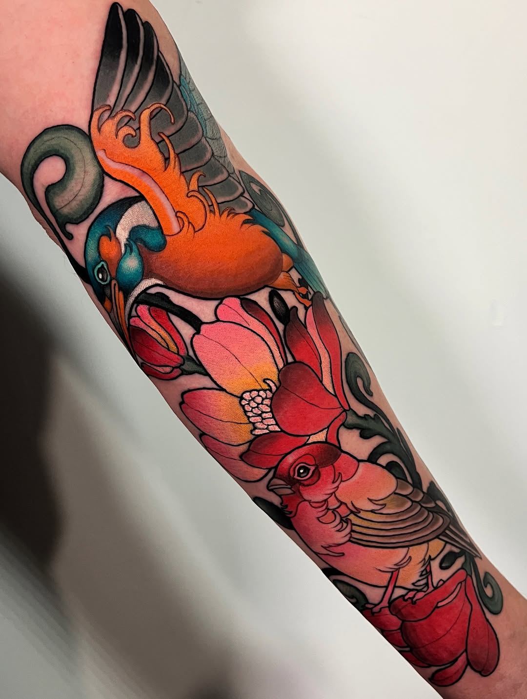 Colorful sleeve tattoo featuring a winged heart, flowering lotus, and bird with orange, red, teal, and black details on the forearm.