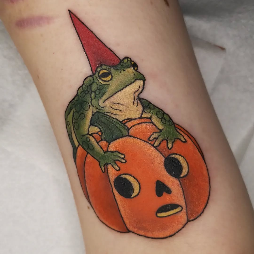 Colorful tattoo on an arm featuring a green toad wearing a red pointed hat, sitting on an orange pumpkin with a face.
