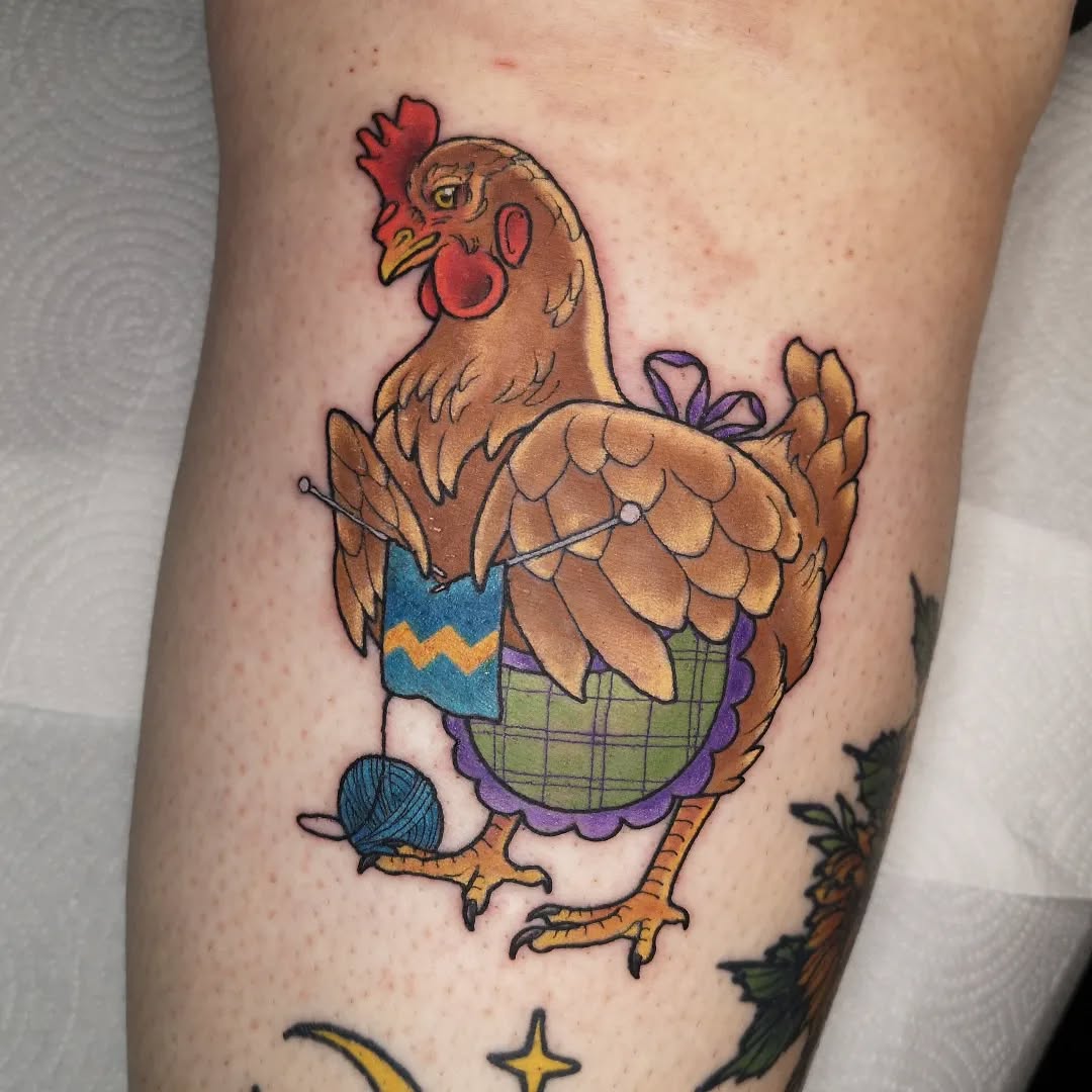 Colorful hen tattoo on leg wearing a blue and yellow sweater and holding knitting yarn, with purple ruffles and orange feet.