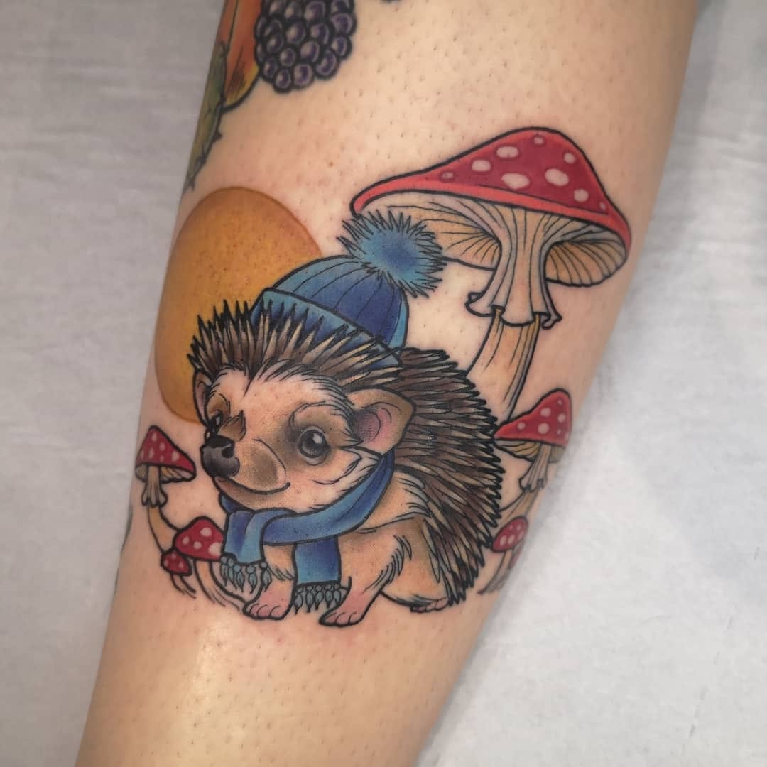 Colorful leg tattoo featuring a hedgehog wearing a blue hat and scarf surrounded by red and white spotted mushrooms and an orange circle.
