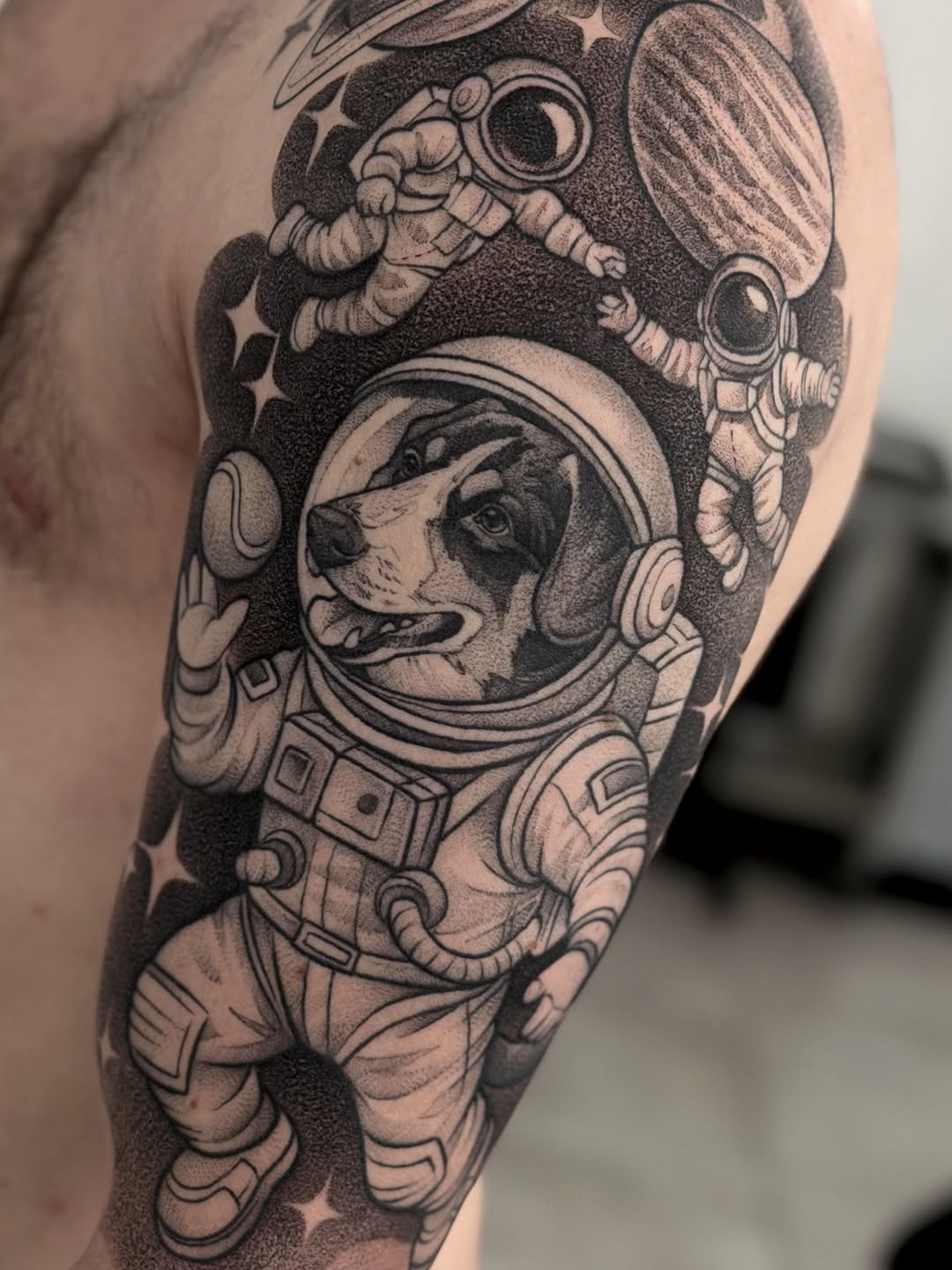 Dog astronaut in spacesuit with helmet and owl above surrounded by stars and celestial elements, tattooed on upper arm in black and gray ink.