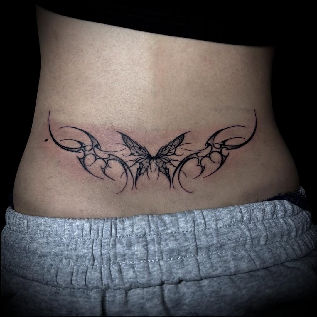 Symmetrical ornamental butterfly tattoo with flowing vine-like wings spanning across the lower back in black ink.