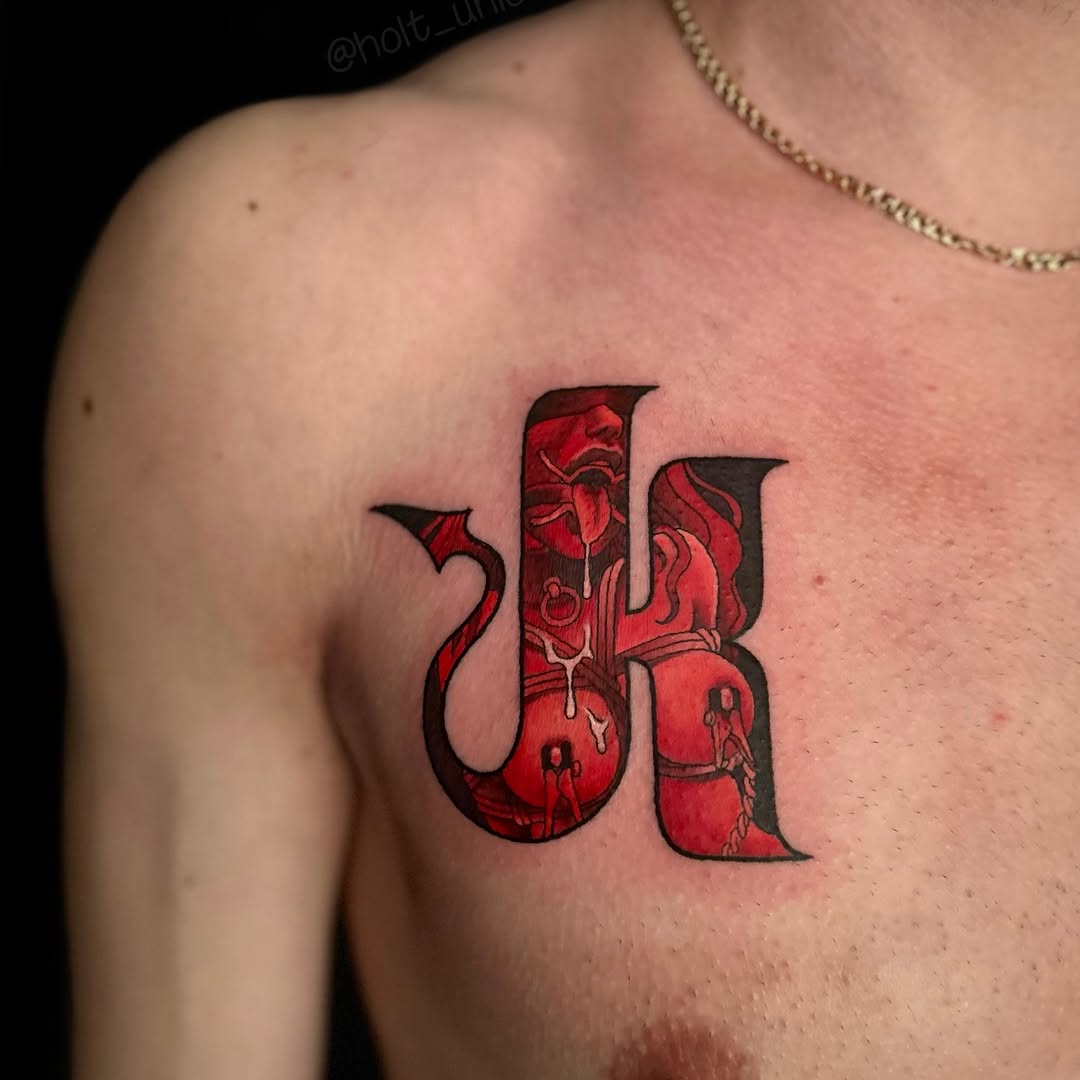Red and black lettering tattoo with floral and figure details on the upper chest of a shirtless person wearing a gold chain.