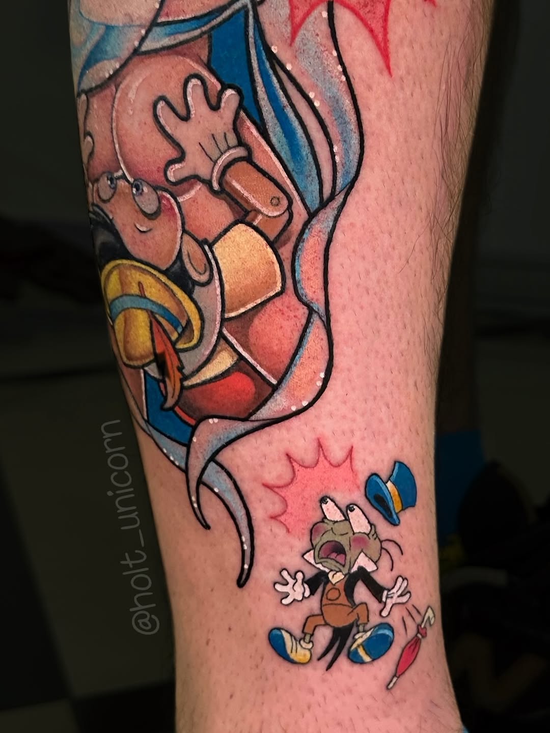 Colorful cartoon tattoo on leg featuring a mermaid with blue tail and a small animated character below, with red, blue, and orange tones.