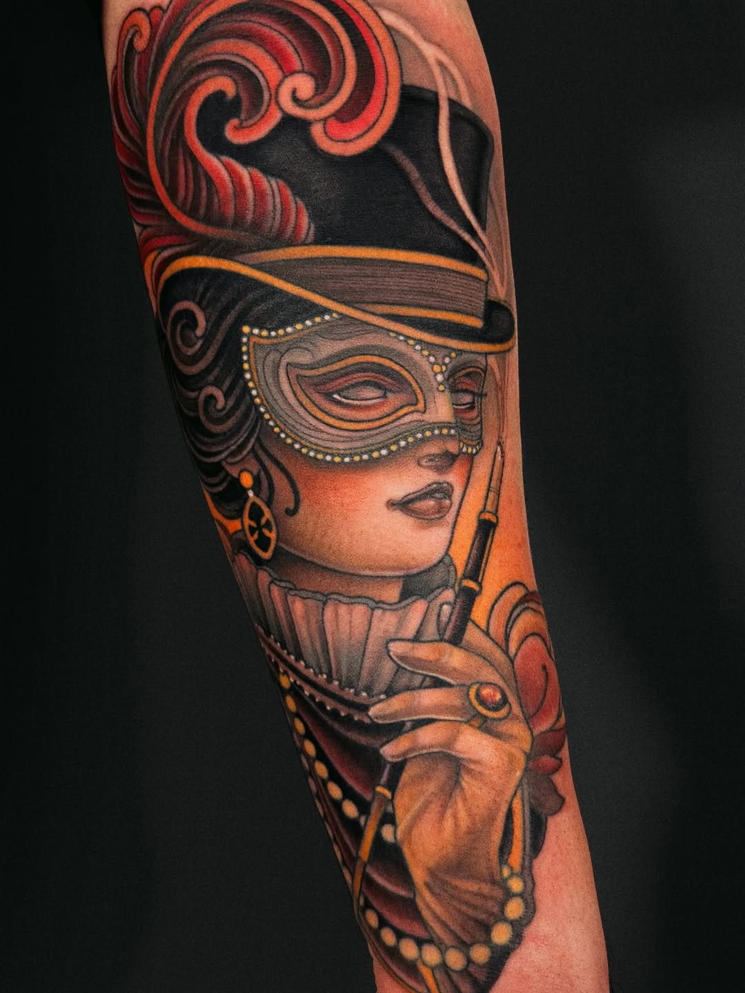 Colorful arm tattoo featuring a masked woman in a decorative hat holding a cigarette holder, with ornate swirls and gold dotted details in orange, red, and black tones.