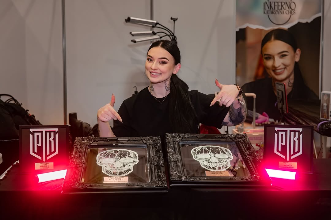 Tattoo artist displays skull design on forearm with black ink during panel event, wearing black clothing and ornate headpiece.