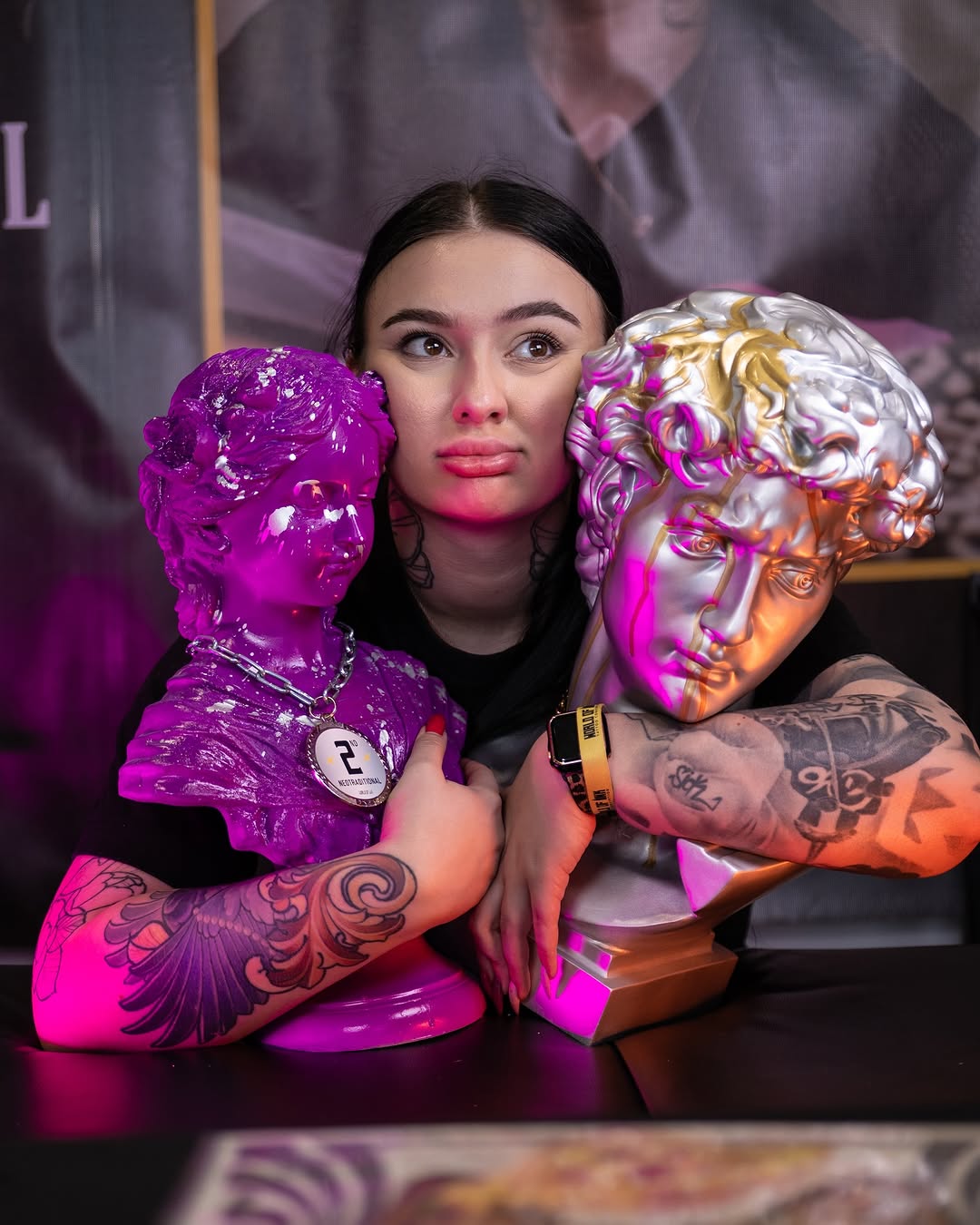 Woman displaying elaborate wing and ornamental tattoos on both arms in purple and orange lighting, posing with decorative sculptural heads.