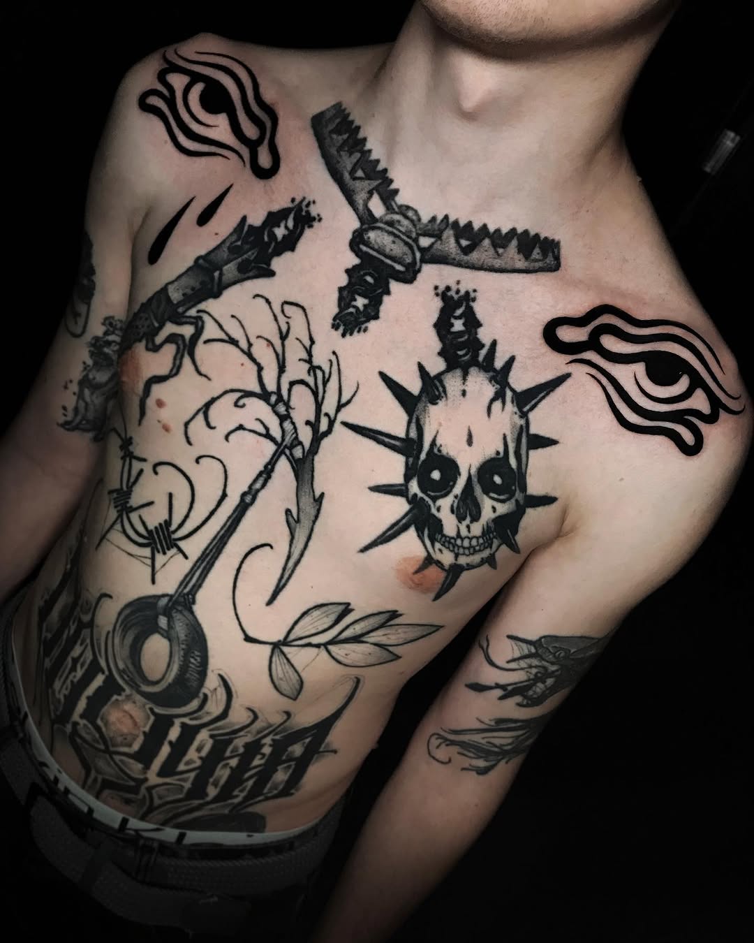 Chest and arm tattoos featuring a spiked skull, birds with outstretched wings, bare tree branches, and decorative line work in black ink.