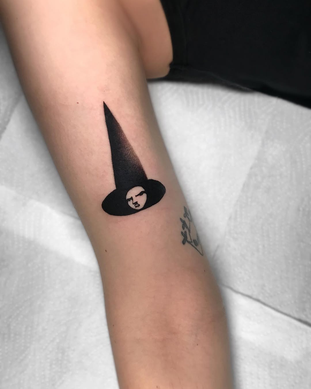 A witch's face wearing a pointed hat with a tall cone extending upward, rendered in solid black ink on the inner forearm.