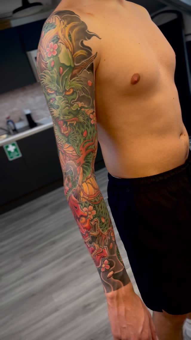 Full-length sleeve tattoo featuring ornate scrollwork, flowers, and decorative elements in green, red, orange, and gold on a person's arm.