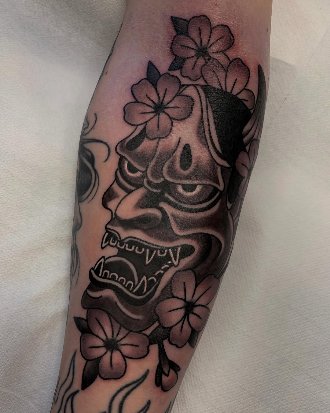 Demon mask with bared teeth and small flowers tattooed on the forearm in black and grey ink with shading.