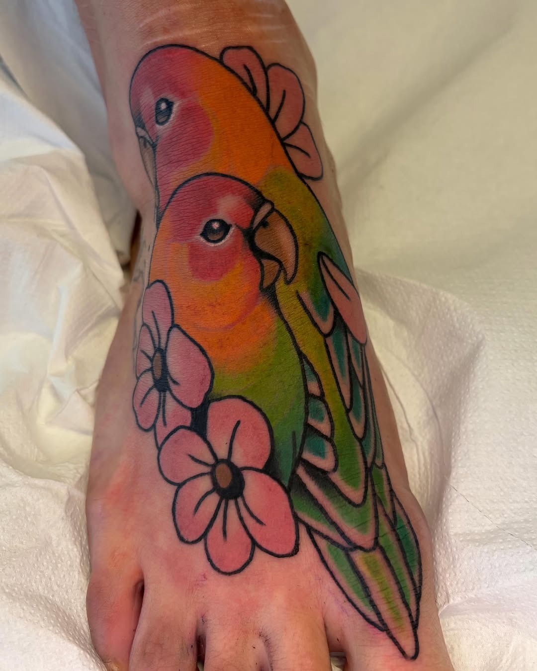 Two colorful parrots with red, orange, and green plumage surrounded by pink flowers tattooed on the back of a hand.