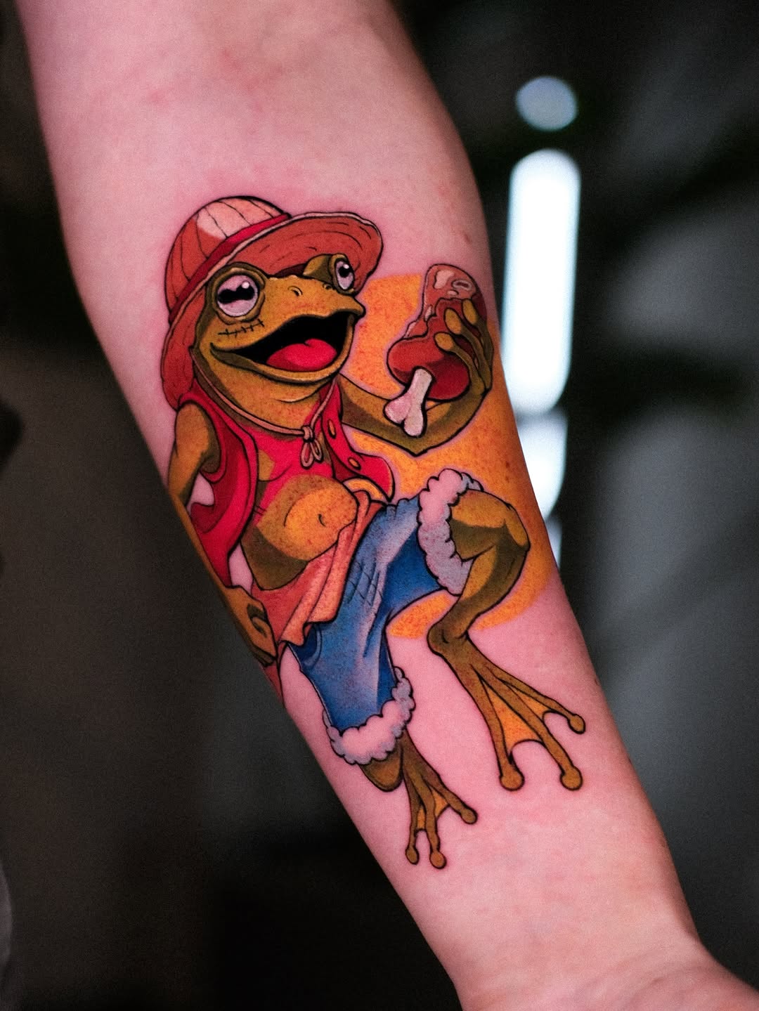 Colorful frog tattoo wearing a red cap and outfit with blue pants, displayed on the inner forearm with orange, red, pink, and blue hues.