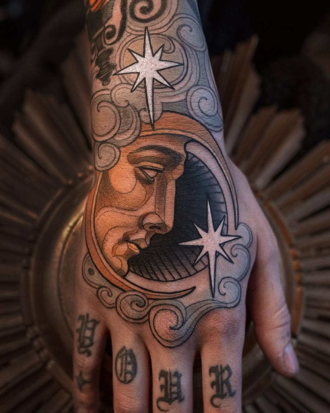 Celestial-themed hand tattoo featuring a crescent moon face with stars, ornamental scrollwork, and additional symbols on the fingers in black and orange ink.