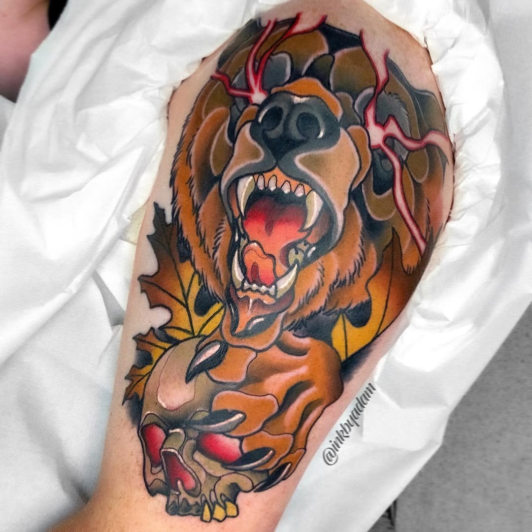 Roaring bear head tattoo on upper arm with orange and brown fur, red mouth, pink veins, yellow claws, and decorative foliage elements.