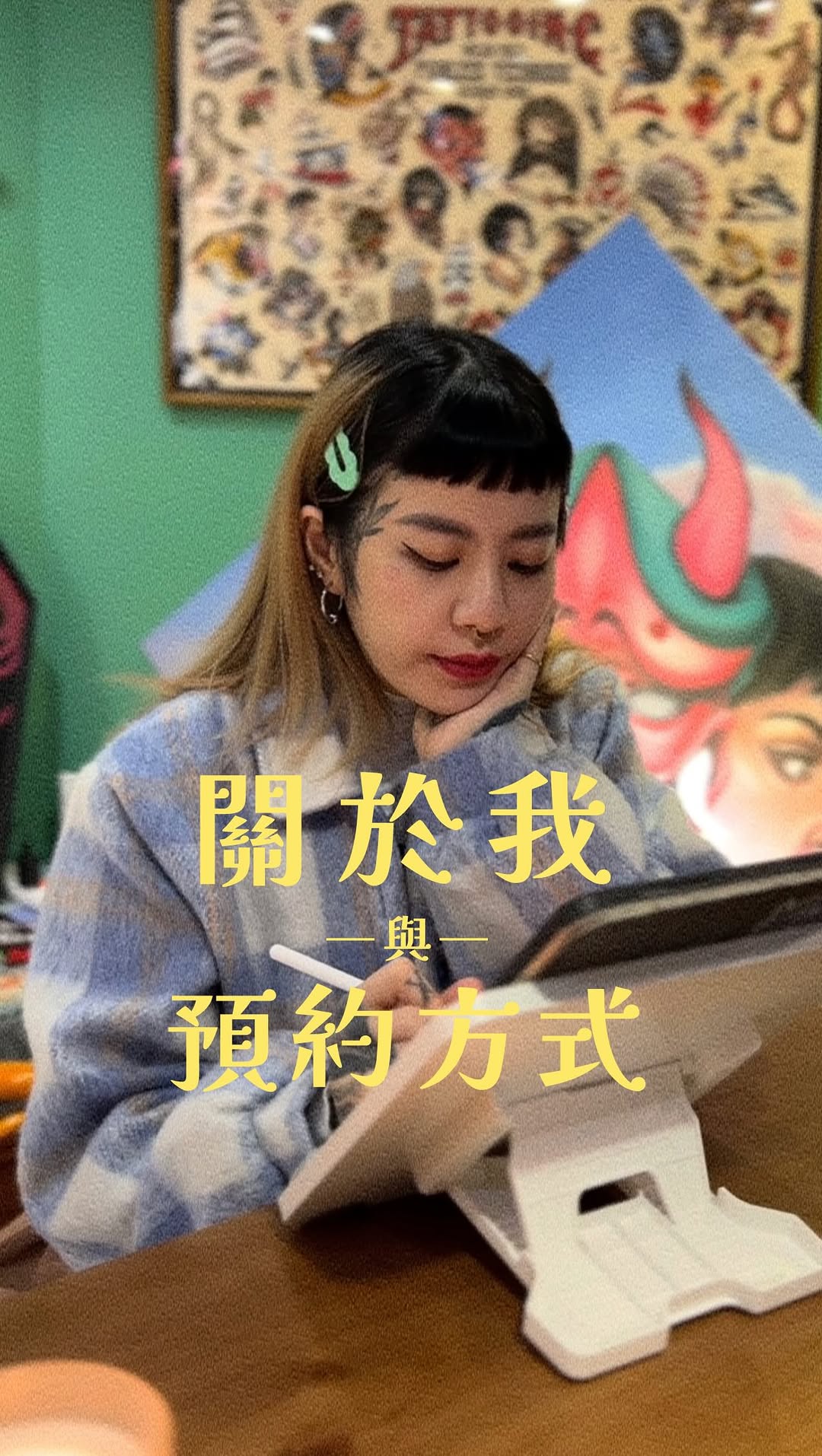 A young woman with a hair clip sits at a table writing, wearing a blue-grey sweater, with colorful artwork visible on green walls behind her.