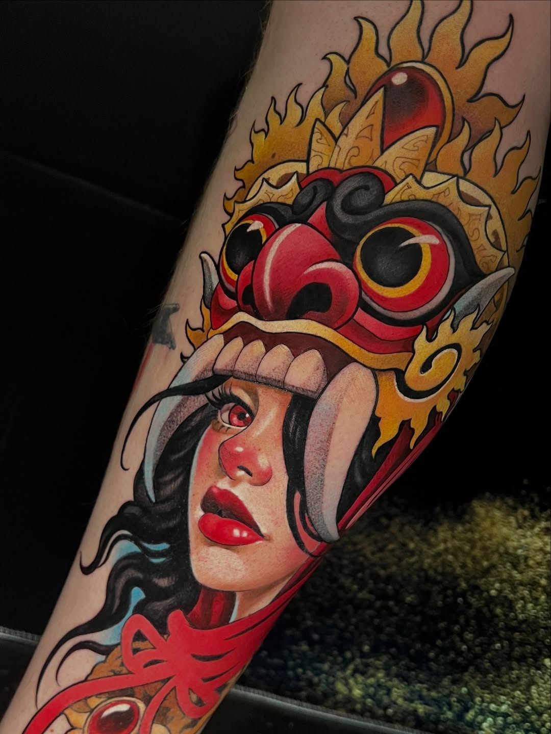 Colorful tattoo on an arm featuring a woman's face with red lips wearing a headdress topped with a red and black mask surrounded by golden flames.