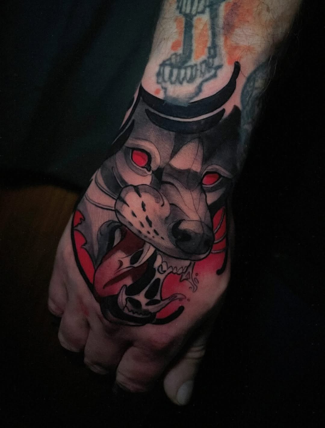 Demon face tattoo with red eyes and black markings on the back of a hand, featuring horns and sharp teeth details.