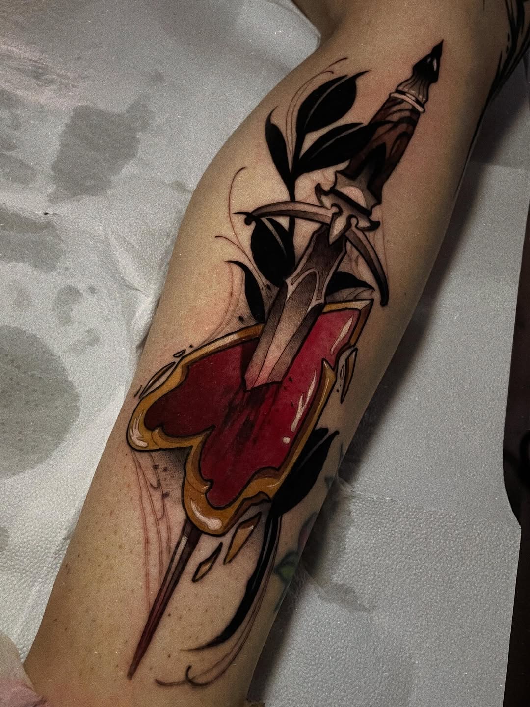 Heart with an arrow through it and decorative leaves on the forearm, featuring red and gold coloring with black line work.